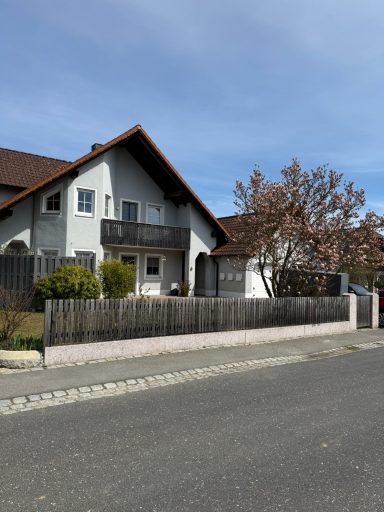 Haus mit Balkon und Garten, umgeben von Bäumen und einem Pflasterweg.
