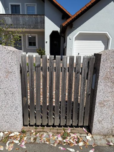 Holzgittertor mit Steinen und Blütenblättern, im Hintergrund ein Haus.