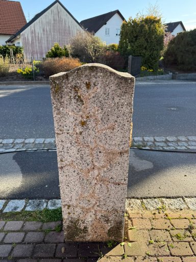 Alter, vermooseter Wegweisungsstein mit unleserlicher Beschriftung in einer Wohnstraße.